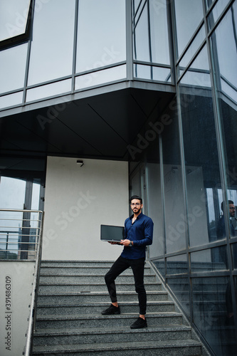 Middle eastern entrepreneur wear blue shirt  eyeglasses with laptop against office building.