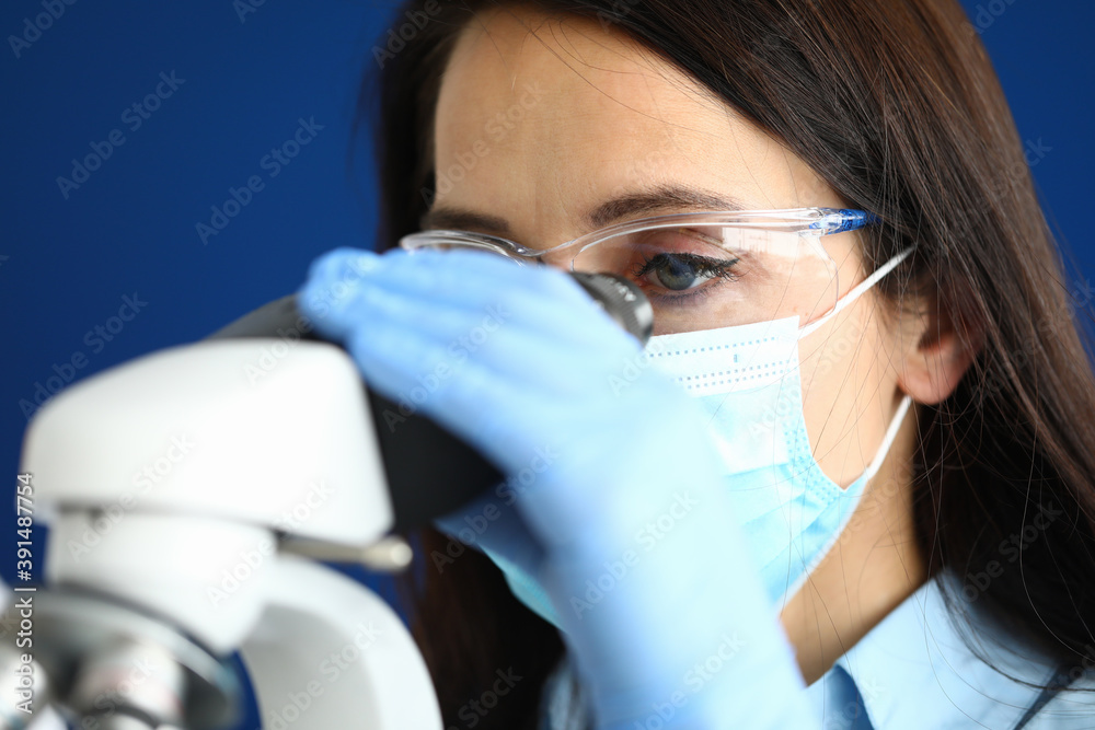 Woman in protective medical mask with glasses and gloves looks through a microscope. New coronavirus research concept