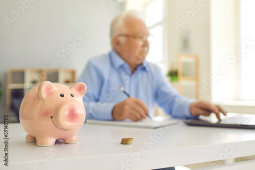 Piggy bank standing on desk with blurred retired man using laptop and doing accounting in background photo