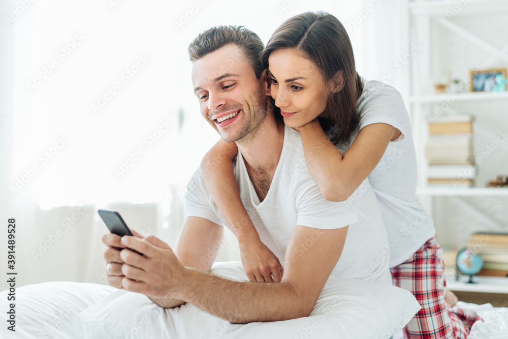 Smiling young beautiful couple using smartphone in bed in sunny morning spending day-off together at home Smiling young beautiful couple using smartphone in bed in sunny morning spending day-off together at home