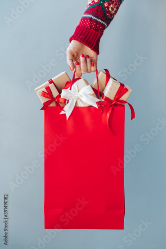 Cropped image of female hand with red polished nails holding plain red shopping bag full of christmas gift boxes