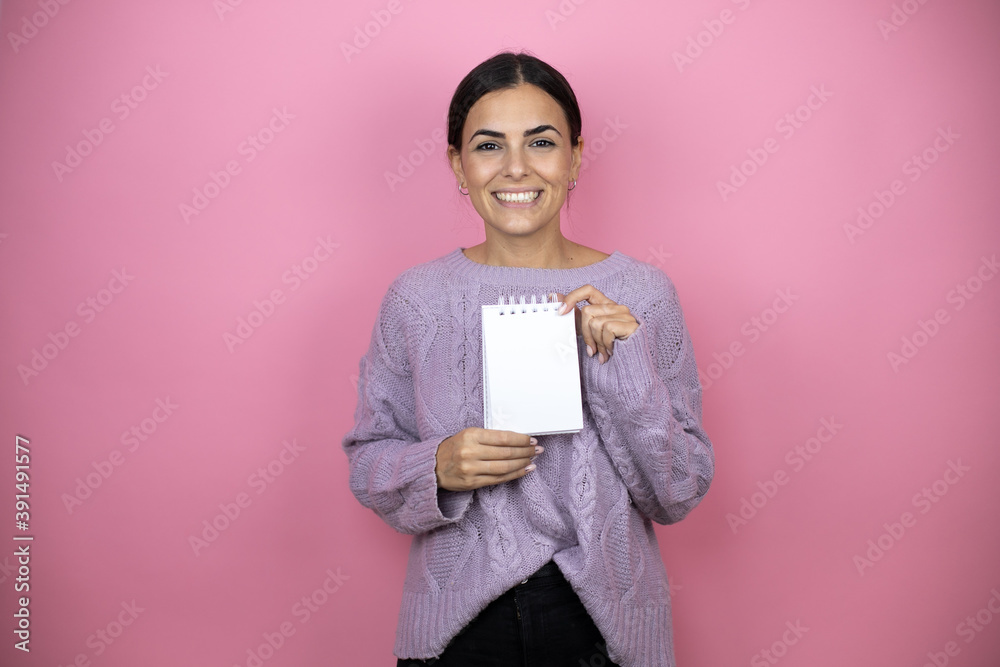 Beautiful woman wearing a casual violet sweater over pink background smiling and showing blank notebook in her hand Beautiful woman wearing a casual violet sweater over pink background smiling and showing blank notebook in her hand