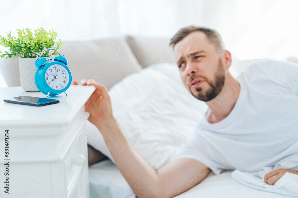 Portrait of a young bearded man looking at his alarm clock unhappy to wake-up and leave the bed Portrait of a young bearded man looking at his alarm clock unhappy to wake-up and leave the bed