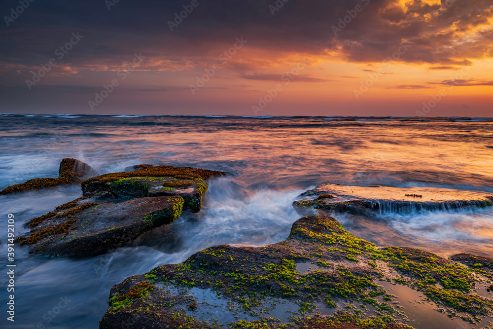 Beautiful seascape. Beach with stones covered by seaweeds. Low tide. Composition of nature. Motion water. Cloudy sky with sunlight. Slow shutter speed. Mengening beach, Bali, Indonesia