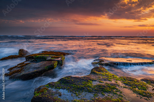 Beautiful seascape. Beach with stones covered by seaweeds. Low tide. Composition of nature. Motion water. Cloudy sky with sunlight. Slow shutter speed. Mengening beach, Bali, Indonesia