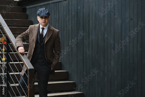 A man posing in the image of an English retro gangster of the 1920s dressed in a coat, suit and flat cap in Peaky blinders style.