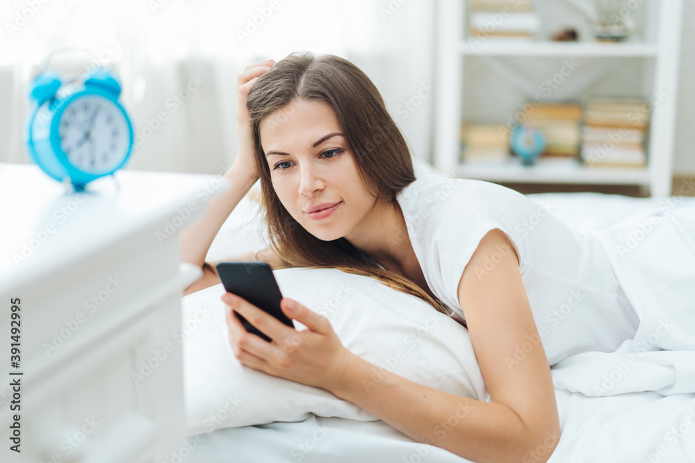 Portrait of attractive young woman lying in bed in the morning and checking message on her smartphone after waking up Portrait of attractive young woman lying in bed in the morning and checking message on her smartphone after waking up