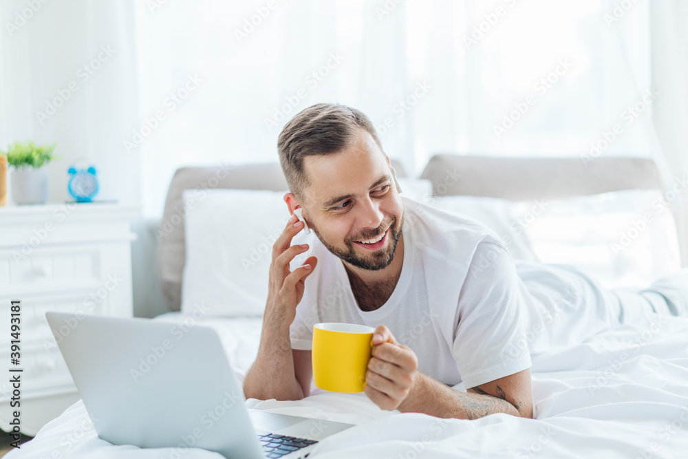 Attractive bearded young man with perfect smile drinking coffee in bed after waking-up and listening to music spending day-off at home