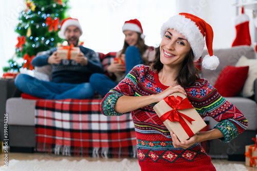 Beautiful smiling young woman in Santa s hat and red sweater with Christmas ornament posing with a gift box with a couple of friends sitting on sofa on the background