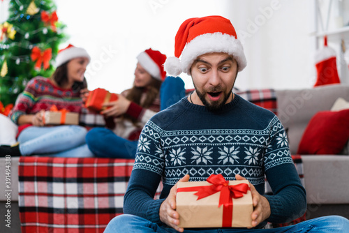 Happy excited young man in Santa Clause hat sitting on a floor and looking with surprised face expression at the gift box in his hands