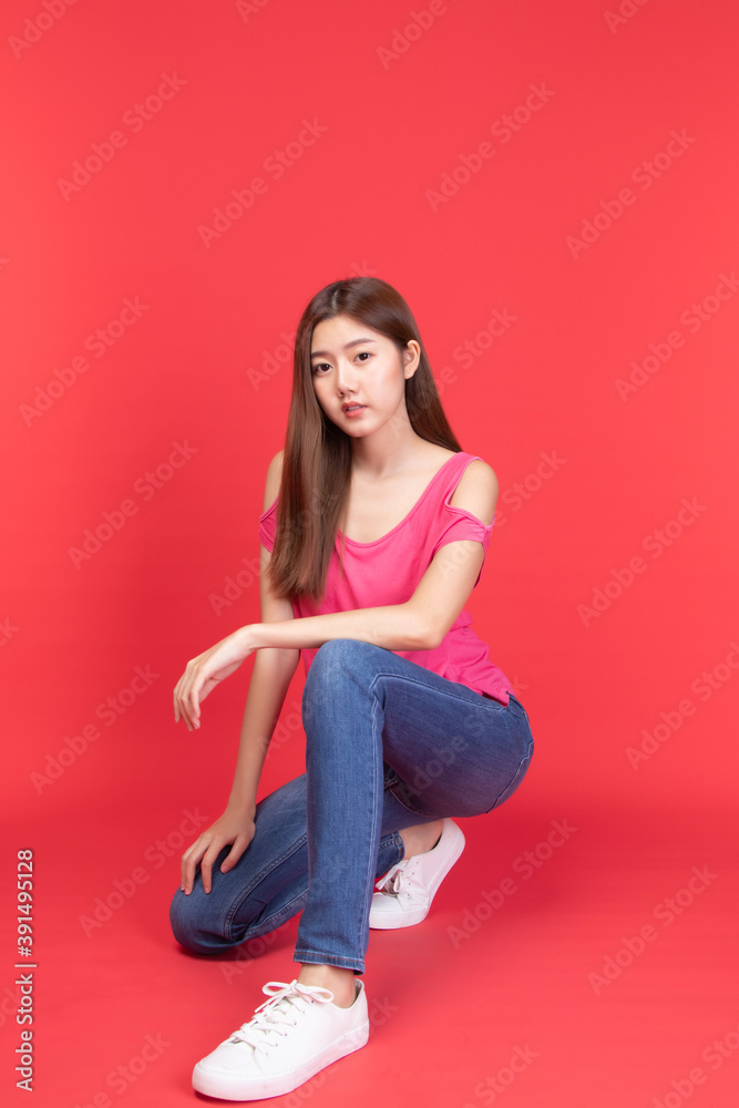 Fashionable young asian model woman in fashion wearing casual red t-shirt with jeans and sneakers sitting on floor in studio on red background. Fashion relax and beauty style. Fashionable young asian model woman in fashion wearing casual red t-shirt with jeans and sneakers sitting on floor in studio on red background. Fashion relax and beauty style.