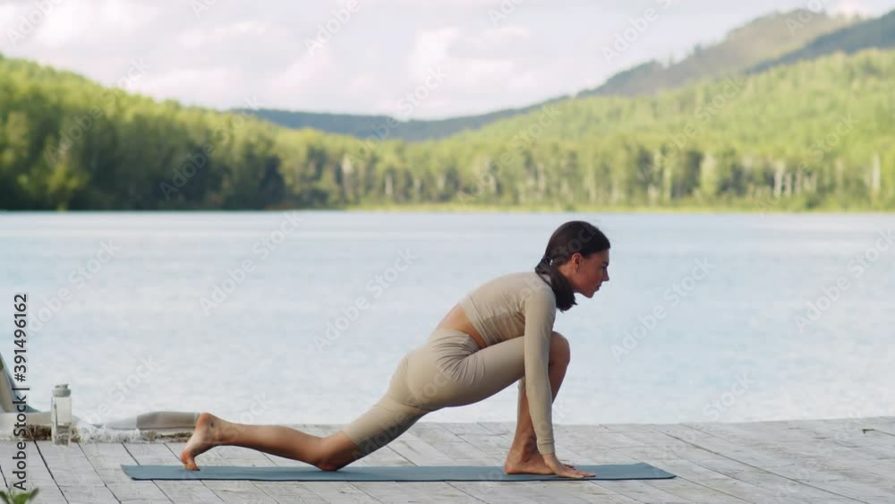 Young fit woman in sportswear standing in forward bend and then doing low lunge with palms together in front of chest while having yoga workout on pier near beautiful lake Young fit woman in sportswear standing in forward bend and then doing low lunge with palms together in front of chest while having yoga workout on pier near beautiful lake