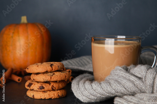 Glass mug of coffee with milk, grey woolen scarf, cookies, spices, orange pumpkins in black table against dark blue wall background. Fall, spicy latte, thanksgiving, coffee shop menu, closeup