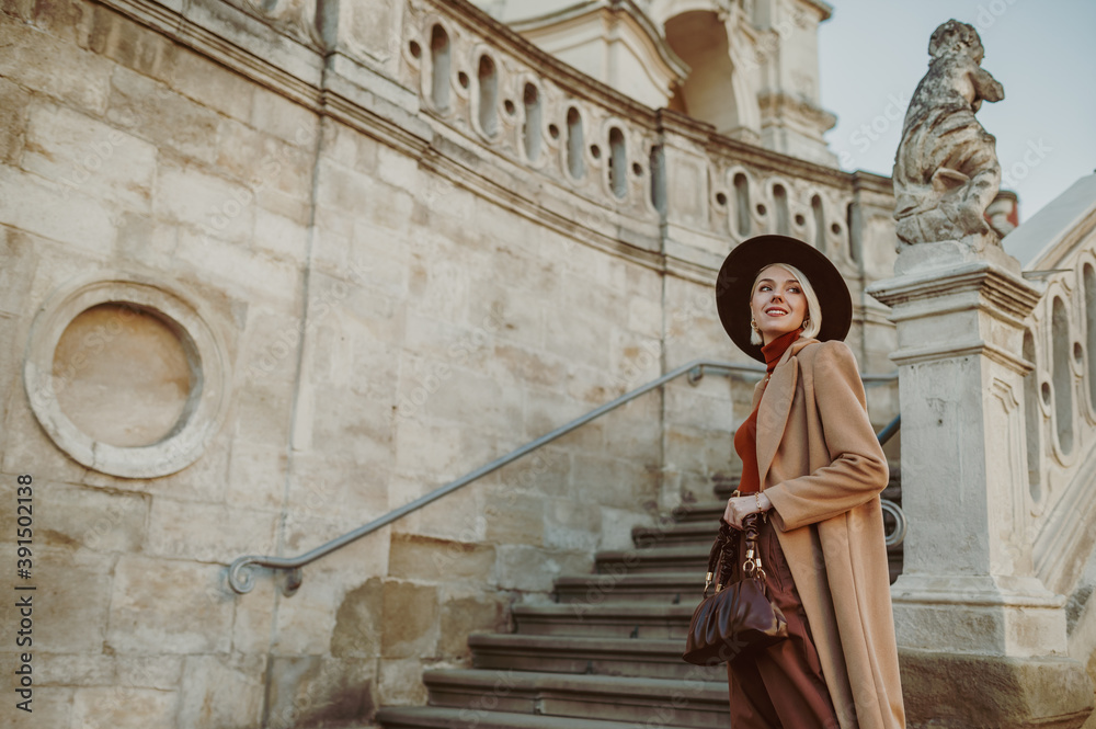 Outdoor autumn fashion portrait of elegant happy smiling woman wearing trendy midi beige, camel color coat, brown hat, holding leather pouch handbag, posing in street of city. Copy, empty space 