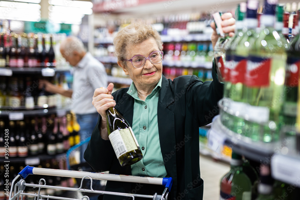 mature woman with glasses chooses bottle of wine in alcohol section of supermarket