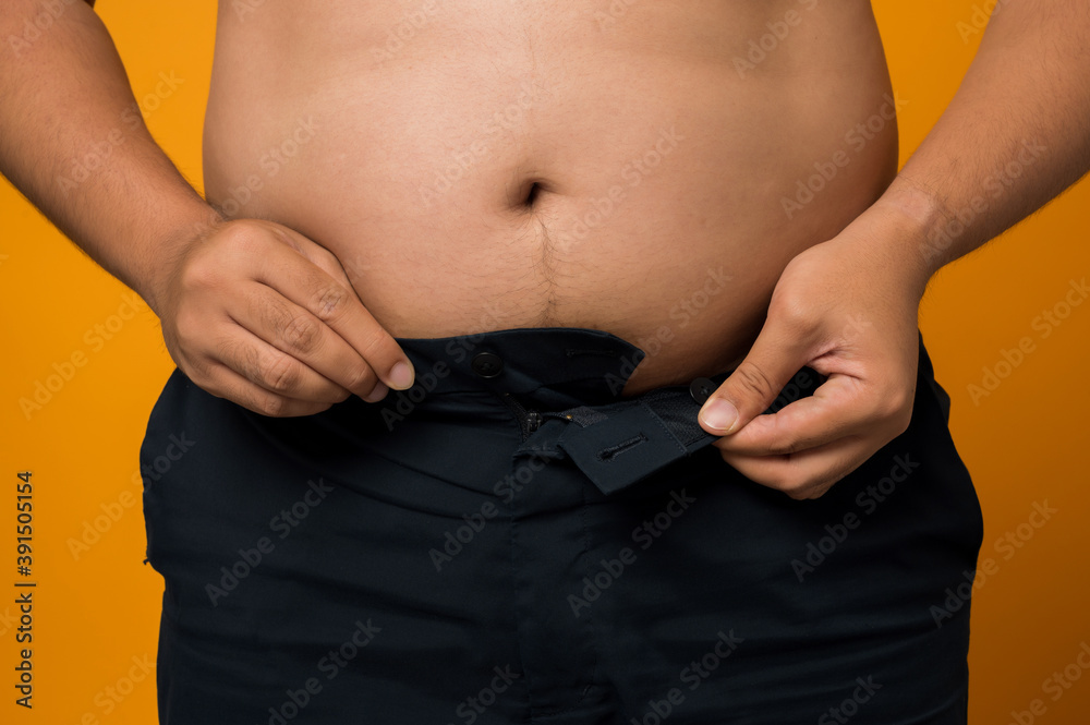 Naklejka premium Close up of overweight man trying to wear too small jeans isolated on yellow background.
