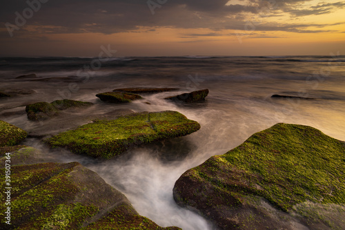 Calm ocean long exposure. Stones covered by green moss in mysterious mist of the sea waves. Concept of nature background. Sunset scenery background. Mengening beach, Bali, Indonesia. © Olga
