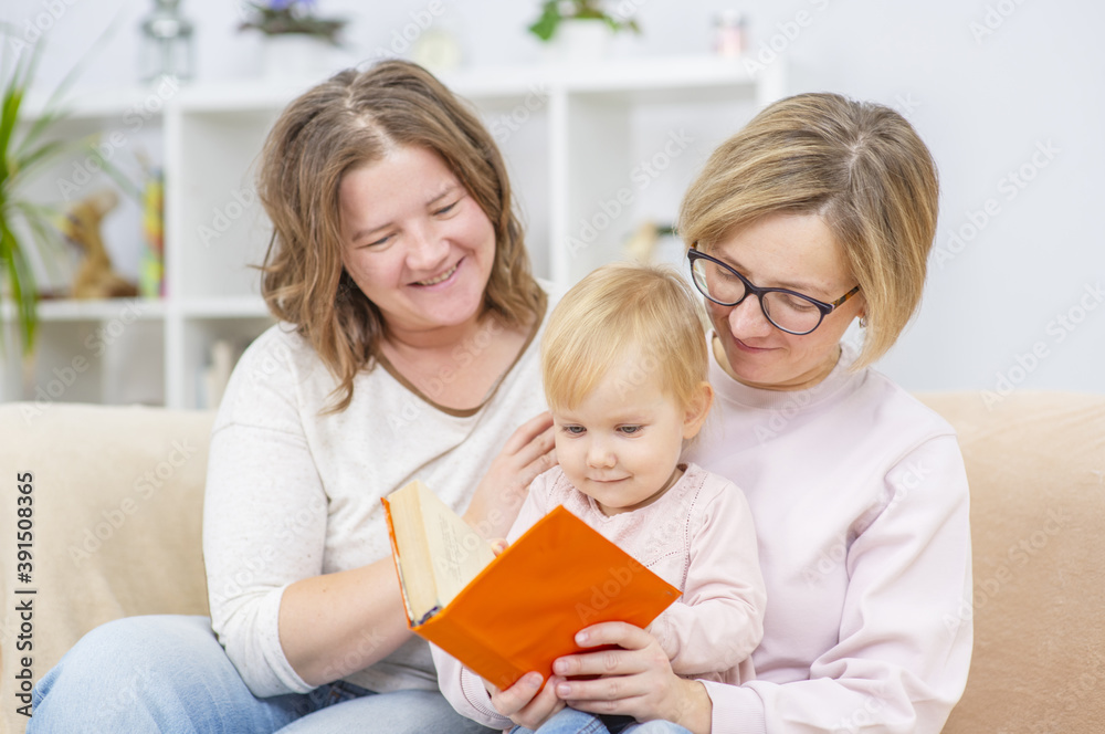 A homosexual family is reading a book to their child. Two girls and a baby are sitting at home on the sofa A homosexual family is reading a book to their child. Two girls and a baby are sitting at home on the sofa