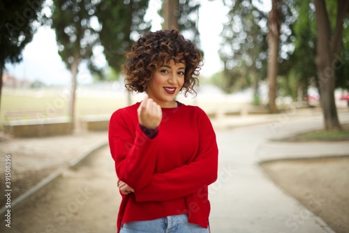 Young arab woman wearing casual red sweater in the street, Beckoning come here gesture with hand inviting welcoming happy and smiling