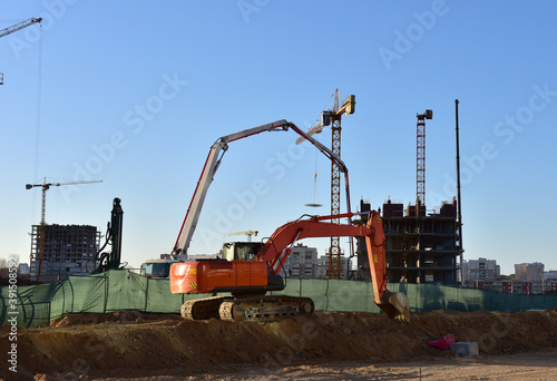 Excavator on earthworks at construction site. Backhoe on foundation work and road construction. Tower cranes in action on blue sky background. Heavy machinery and construction equipment