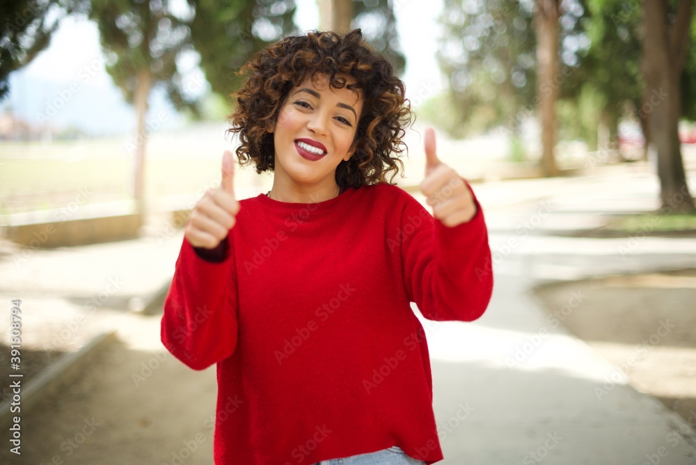 Young arab woman wearing casual red sweater in the street, approving doing positive gesture with hand, thumbs up smiling and happy for success. Winner gesture.