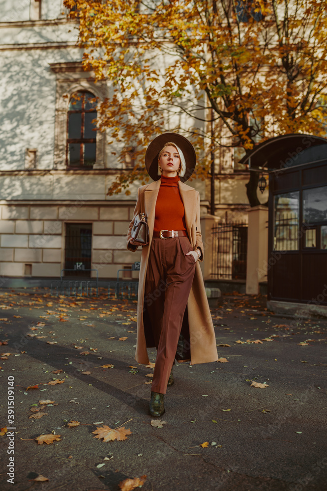 Outdoor full-length autumn fashion portrait of elegant, luxury woman wearing trendy long, midi beige, camel color coat, brown hat, classic trousers, orange turtleneck, walking in street of city
