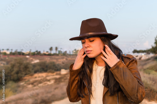 Young woman with hat and brown leather jacket, straightens her hair, in natural park. © Marcela Ruty Romero