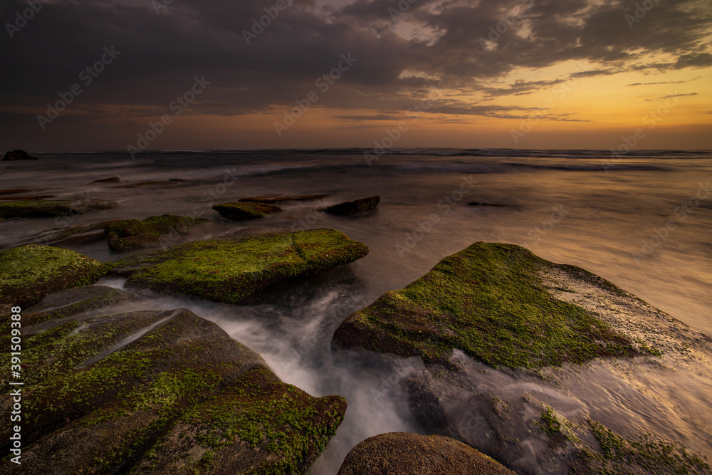 Calm ocean long exposure. Stones covered by green moss in mysterious mist of the sea waves. Concept of nature background. Sunset scenery background. Mengening beach, Bali, Indonesia.