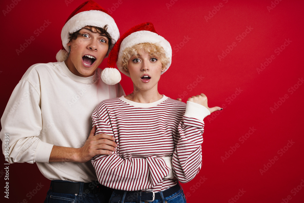 Fototapeta premium Young shocked couple in Santa Claus hats pointing finger aside