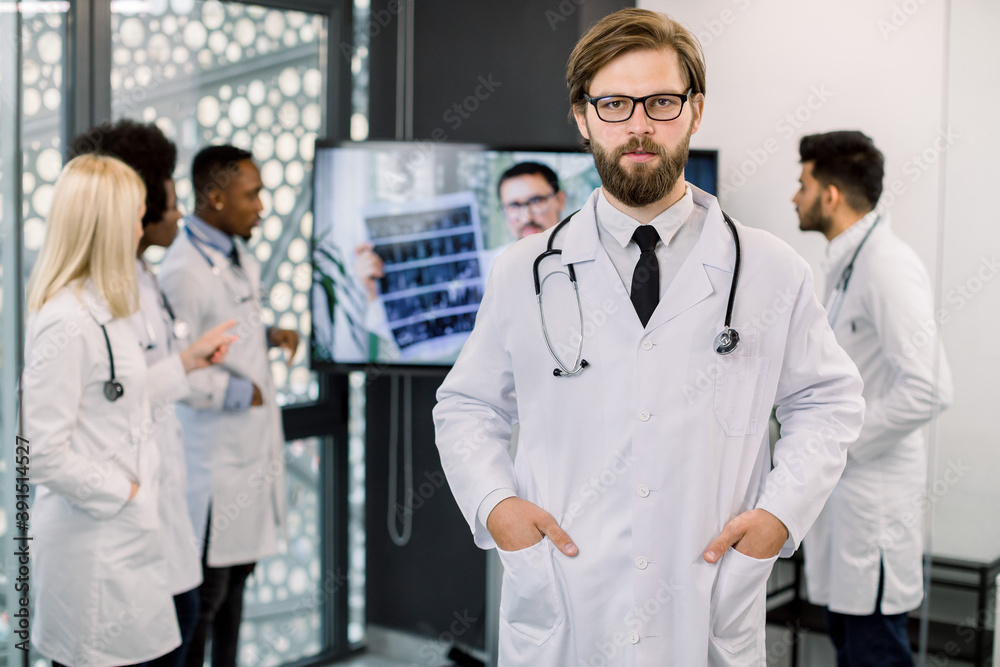 Handsome young bearded Caucasian male doctor posing to camera at modern clinic. Team of multi-ethnic doctors on the background, discussing CT results and having video call with male doctor Handsome young bearded Caucasian male doctor posing to camera at modern clinic. Team of multi-ethnic doctors on the background, discussing CT results and having video call with male doctor
