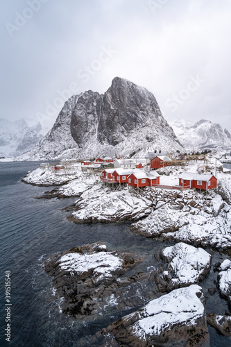 a small serene Norwegian fishing village on a shore in the north. The town, big mountain, and cliff covered with white fluffy snow in the beautiful winter under overcast sky. Waves break on the rocks.