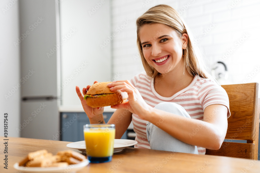 Smiling young woman eating hamburger at the kitchen