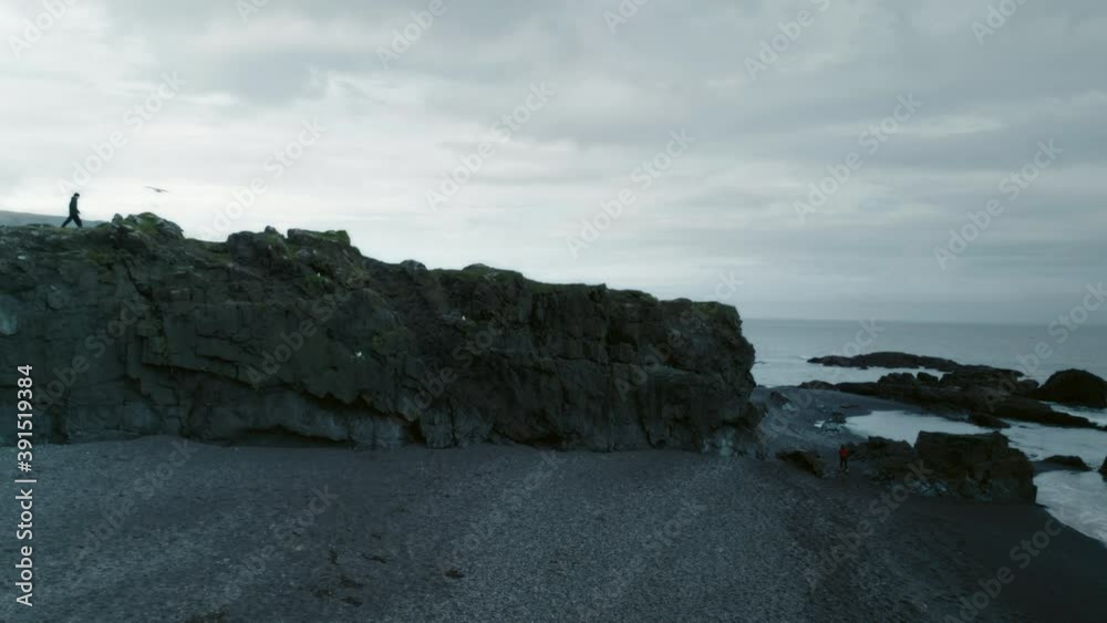 Silhouette of lonely man walk on edge of cliff rocks at moody gloomy beach before storm. Atmospheric shot of grey and dark sky on volcanic icelandic beach Silhouette of lonely man walk on edge of cliff rocks at moody gloomy beach before storm. Atmospheric shot of grey and dark sky on volcanic icelandic beach