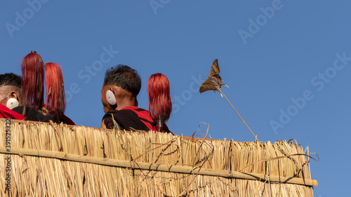 Naga men siting with only their heads and their traditional weapons visible behind thatched fence and a butterfly stuck in a twig at a village at kohima Nagaland India photo
