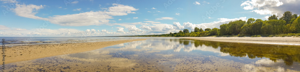 Calm sea on a sunny summer day. Blue cloudy sky and tree reflection in tranquil water. Holiday and travel concept. Latvian nature. Panoramic view.