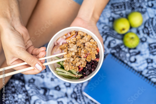High angle of woman having picnic outdoor sitting on blue blanket with chopsticks eating takeout healthy vegetarian poke bowl with tofu, fermented soybeans -tempeh, cucumber, rice, carrots, onion
