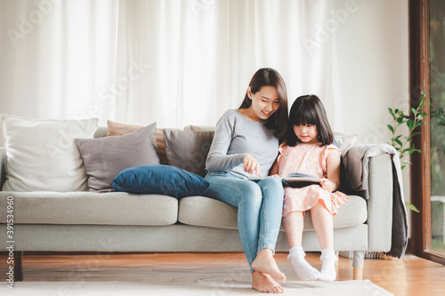 Happy Asian family mother and daughter sitting on sofa reading a book in living room at home