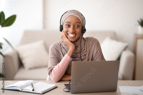 Portrait of beautiful Muslim black woman in headscarf wearing headset, communicating online on laptop computer, indoors photo