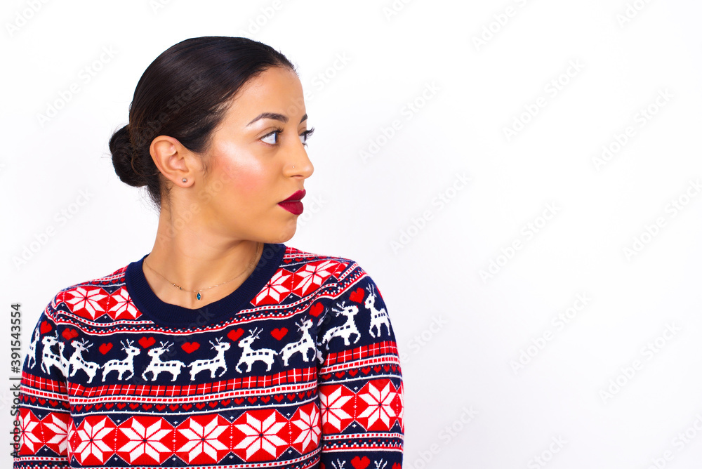 Side view of young happy smiling Young beautiful Arab woman wearing Christmas sweater against white wall Side view of young happy smiling Young beautiful Arab woman wearing Christmas sweater against white wall