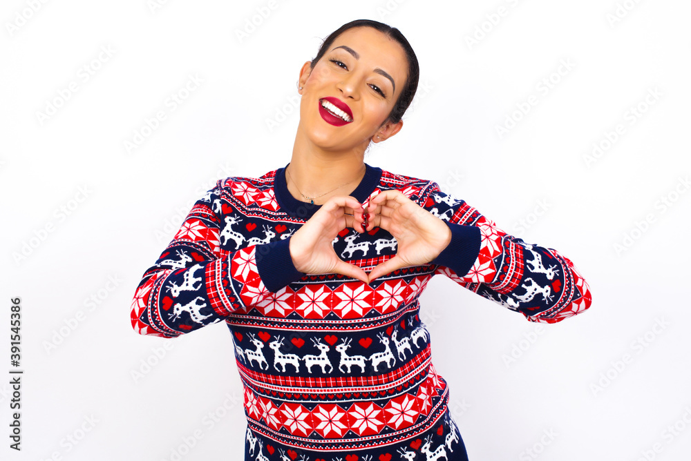 Young beautiful Arab woman wearing Christmas sweater against white wall smiling in love showing heart symbol and shape with hands. Romantic concept. Young beautiful Arab woman wearing Christmas sweater against white wall smiling in love showing heart symbol and shape with hands. Romantic concept.