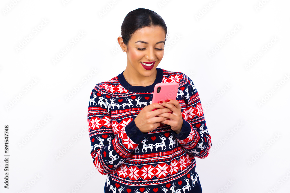 Smiling Young beautiful Arab woman wearing Christmas sweater against white using cell phone, messaging, being happy to text with friends, looking at smartphone. Modern technologies and communication. Smiling Young beautiful Arab woman wearing Christmas sweater against white using cell phone, messaging, being happy to text with friends, looking at smartphone. Modern technologies and communication.
