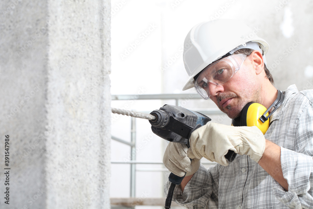 man using an electric pneumatic drill making a hole in wall, professional construction worker with safety hard hat, hearing protection headphones, gloves and protective glasses. - obrazy, fototapety, plakaty 