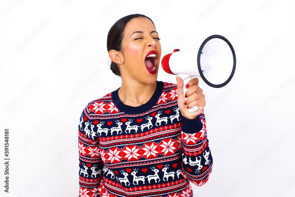 Young beautiful Arab woman wearing Christmas sweater against white wall, communicates shouting loud holding a megaphone, expressing success and positive concept, idea for marketing or sales. Young beautiful Arab woman wearing Christmas sweater against white wall, communicates shouting loud holding a megaphone, expressing success and positive concept, idea for marketing or sales.