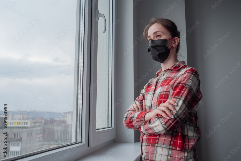 Naklejka premium Portrait of pensive woman wearing black medical face mask, red plaid shirt and looking out of window in room with grey wall at home. Self isolation, quarantine, COVID 19, coronavirus, safety concept
