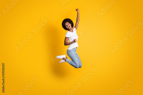 Full length body size photo of cheerful happy girl with black skin jumping high keeping hand up laughing isolated on bright yellow color background