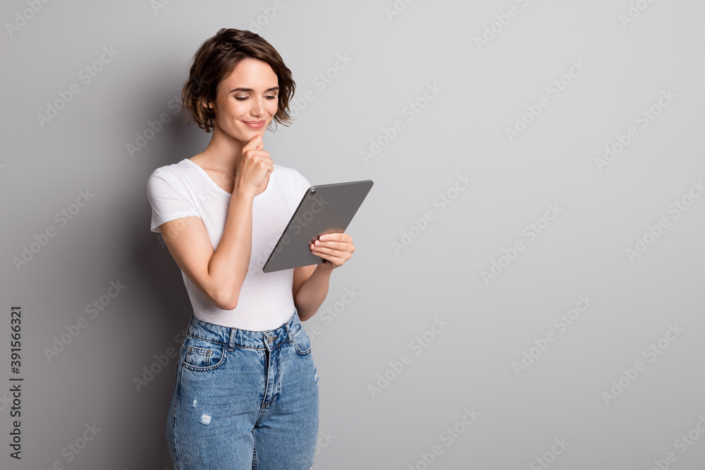 Photo of attractive young girl hold pc tablet read browse news information hand touch chin isolated over grey color background Photo of attractive young girl hold pc tablet read browse news information hand touch chin isolated over grey color background