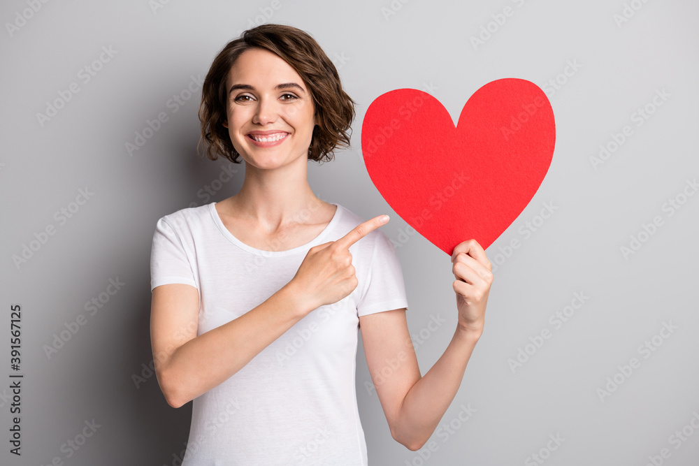 Photo of young happy smile girl point index finger hold big heart valentine day advertise offer isolated over grey color background Photo of young happy smile girl point index finger hold big heart valentine day advertise offer isolated over grey color background
