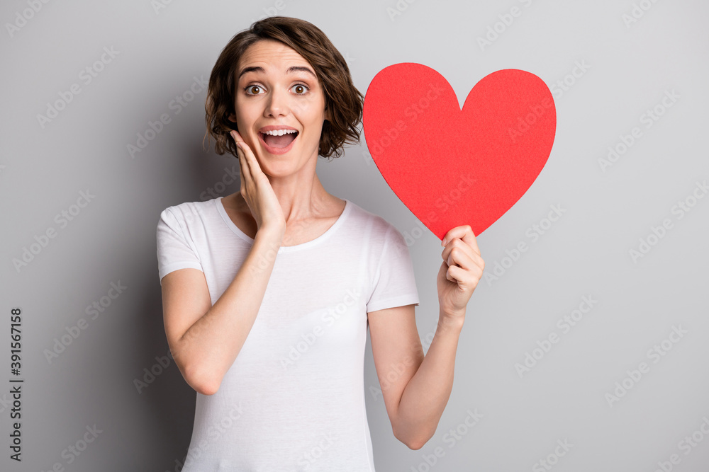 Photo of surprised amazed young girl hand touch cheeks hold paper heart feelings love isolated over grey color background Photo of surprised amazed young girl hand touch cheeks hold paper heart feelings love isolated over grey color background
