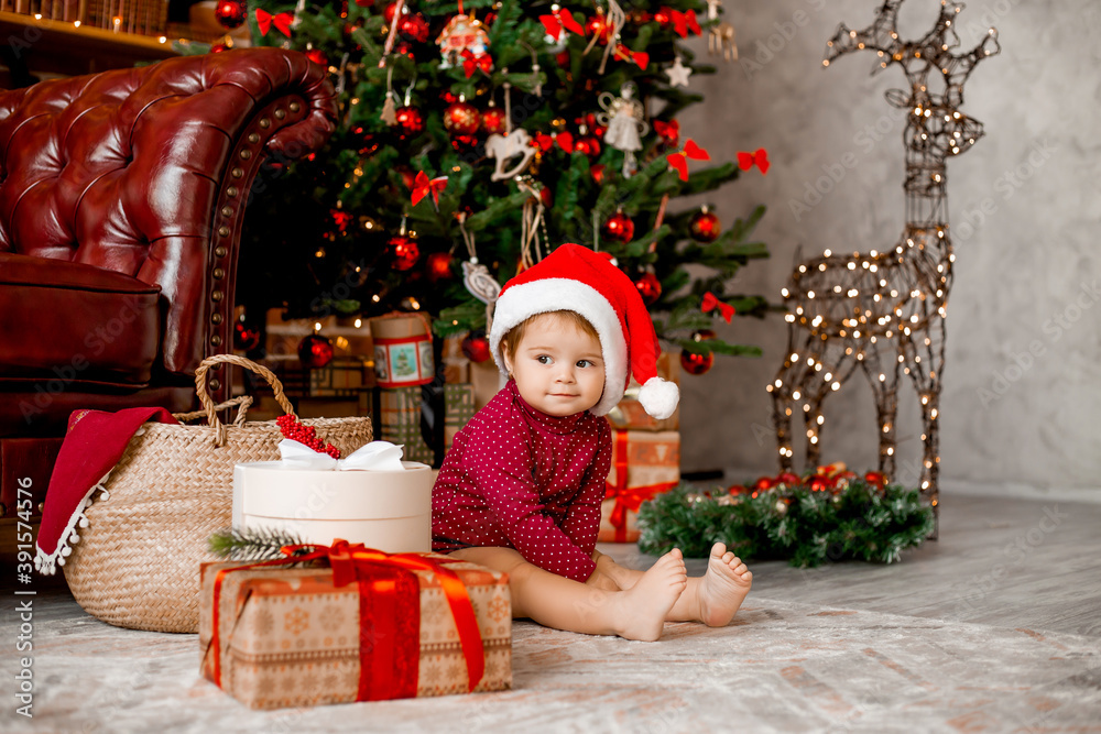Cute baby Santa sits at home near the Christmas tree with gifts