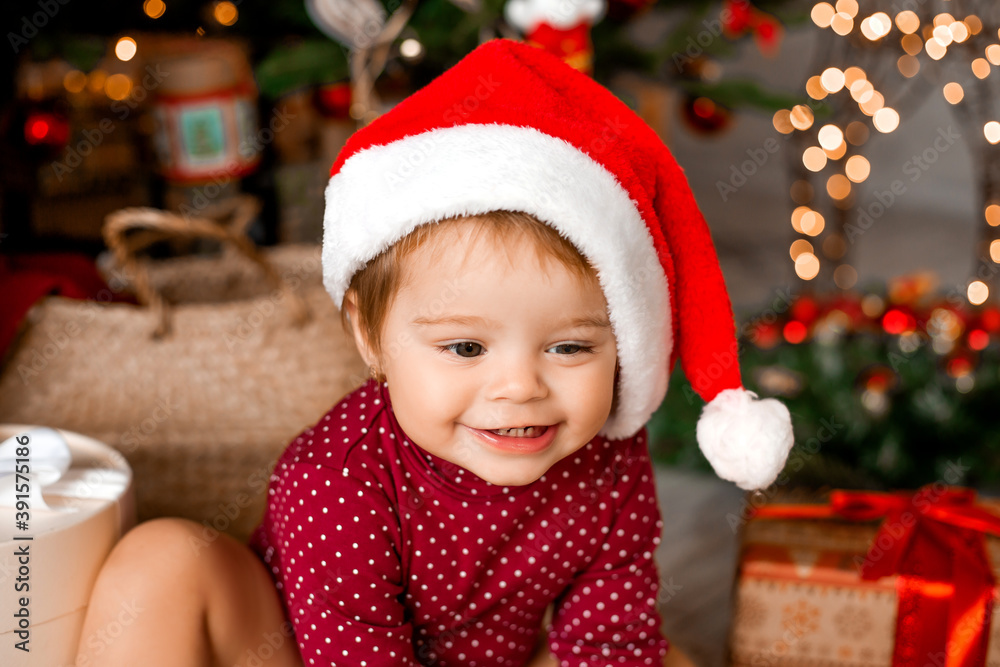 Cute baby Santa sits at home near the Christmas tree with gifts Cute baby Santa sits at home near the Christmas tree with gifts